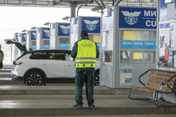 Bulgarian border police officer in high-vis vest inspecting white SUV with open boot at customs checkpoint with migration control booths visible