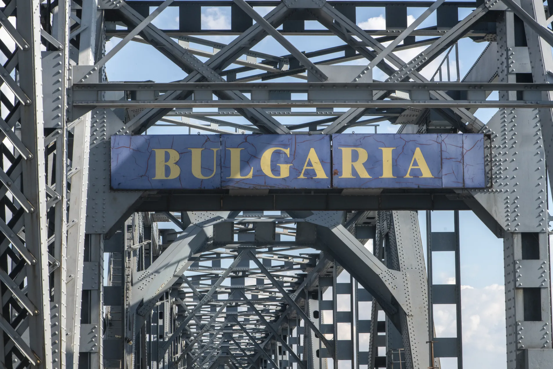 Close-up of weathered gold Bulgaria entry sign mounted on historic steel truss Danube Bridge structure against blue sky